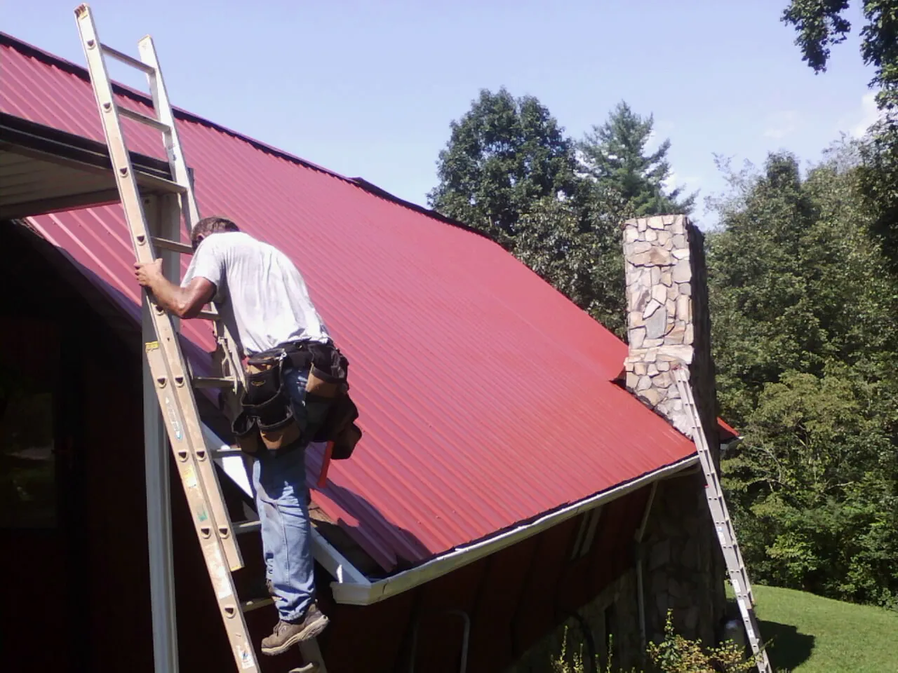 Roofer on a ladder beside a red metal roof and stone chimney — Soffit & Fascia Repair by Roofing Brothers in Hampton Falls